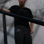 A person wearing a black short-sleeve t-shirt with gold lettering and dark gray wide-leg jeans, standing on a staircase next to a black metal railing against a textured gray wall.